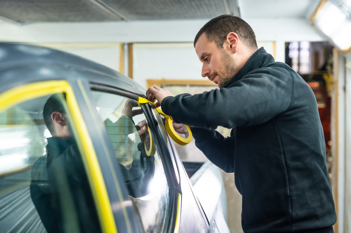 Auto body shop technician applying yellow masking tape to a car door window