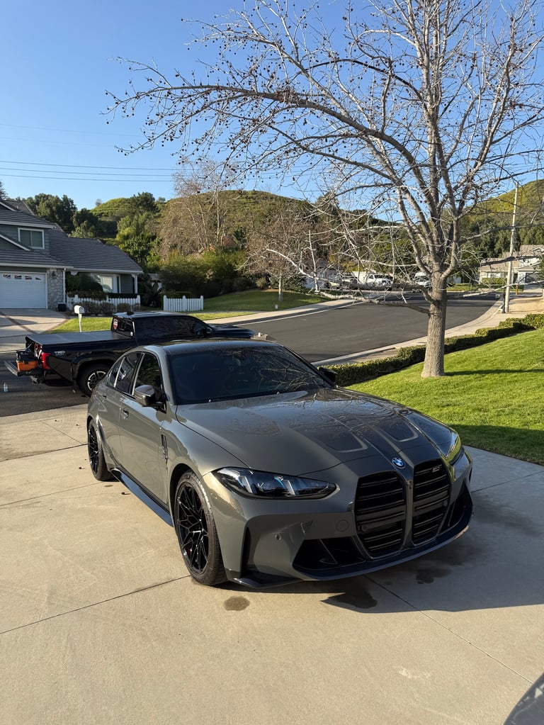 Silver BMW M440i sports sedan parked in residential driveway on sunny day with bare tree and hills in background