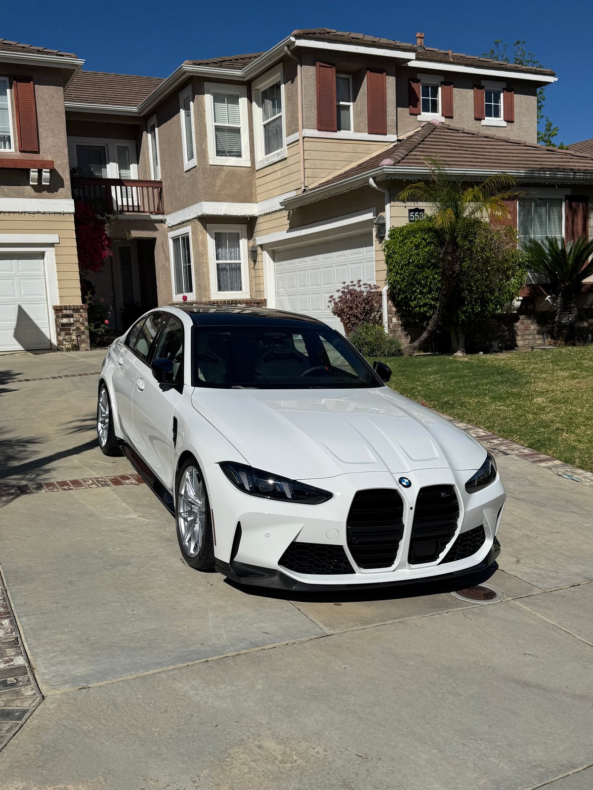 White BMW M340i sedan parked in driveway of beige and brown multi-story house on clear sunny day